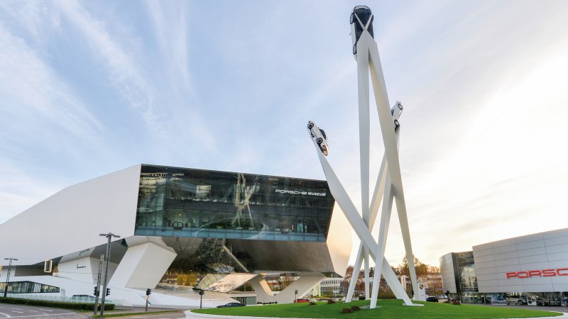 Das Porsche Museum mit seiner modernen Architektur und einer Skulptur von drei Autos auf hohen Säulen vor einem blauen Himmel., © Porsche AG Sabine Braun