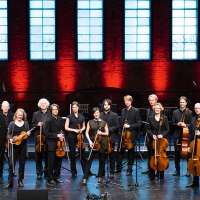 Das Stuttgarter Kammerorchester posiert auf einer B&uuml;hne vor einer roten Backsteinwand mit gro&szlig;en Fenstern. Die Musiker halten ihre Instrumente., &copy; Reiner Pfisterer