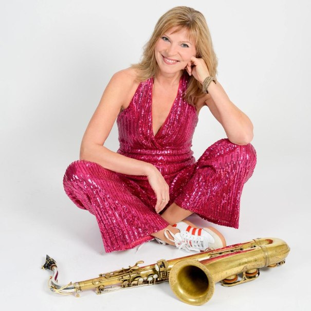 Eine l&auml;chelnde Frau in einem pinken Paillettenkleid sitzt auf dem Boden neben einem Saxophon. Sie tr&auml;gt wei&szlig;e Turnschuhe mit roten Streifen., &copy; Foto Ruth Sabadino