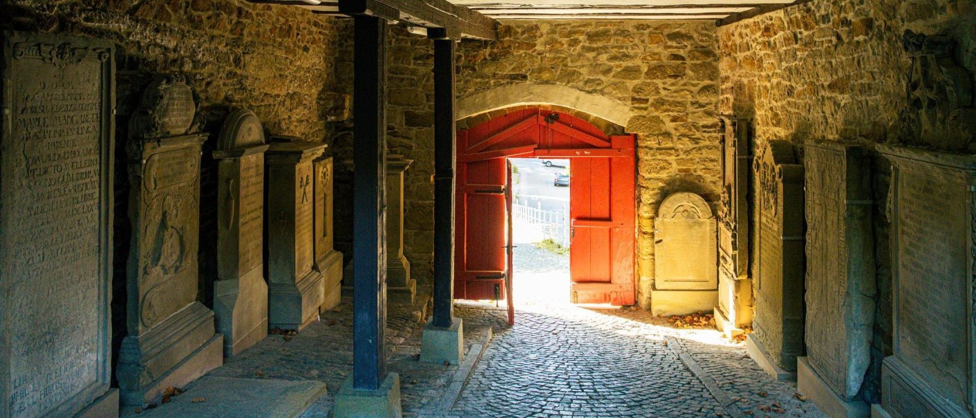 Ein historischer Durchgang mit Grabsteinen an den Wänden und einem roten Tor, das nach draußen führt. Sonnenlicht fällt durch das Tor auf den Boden., © Stuttgart-Marketing GmbH, Sarah Schmid Ein historischer Durchgang mit Grabsteinen an den Wänden und einem roten Tor, das nach draußen führt. Sonnenlicht fällt durch das Tor auf den Boden., © Stuttgart-Marketing GmbH, Sarah Schmid
