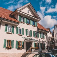 Ein traditionelles Gasthaus mit grünen Fensterläden und roten Dachziegeln, umgeben von anderen Gebäuden. Der Himmel ist blau mit einigen Wolken., © Stadt Welzheim