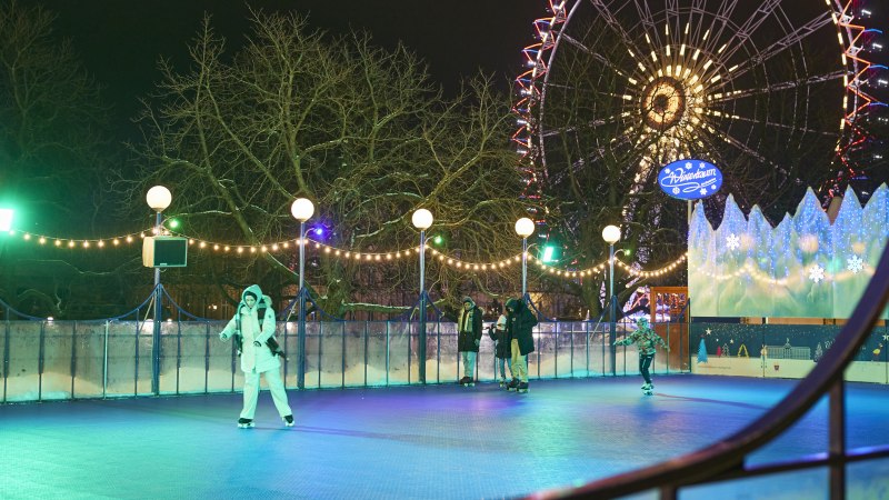 Eislaufbahn bei Nacht, beleuchtet mit Lichterketten. Menschen laufen Schlittschuh, im Hintergrund ein beleuchtetes Riesenrad und ein Schild 'Wintertraum'., © SMG, Sarah Schmid