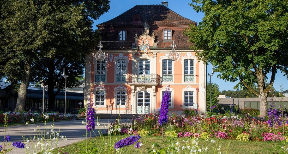 Rokokoschlösschen mit rosa Fassade, verziert mit weißen Ornamenten, umgeben von blühenden Blumen und Bäumen im Sonnenschein., © Mario Klaiber Rokokoschlösschen mit rosa Fassade, verziert mit weißen Ornamenten, umgeben von blühenden Blumen und Bäumen im Sonnenschein., © Mario Klaiber