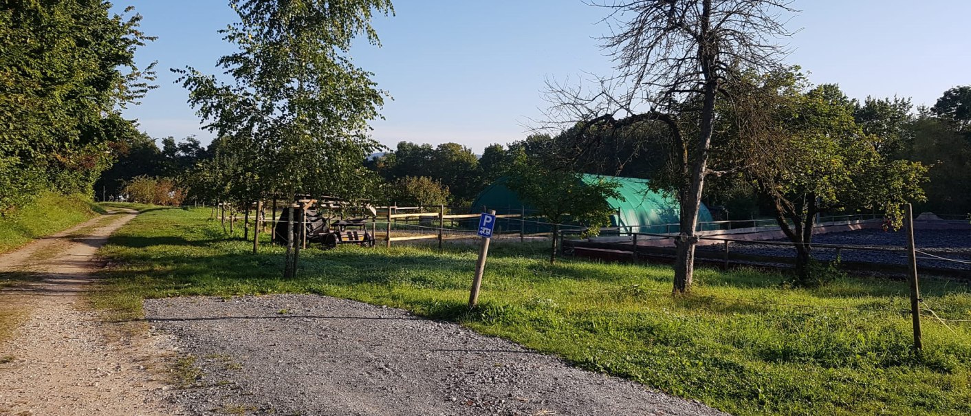 Ein Feldweg führt durch eine grüne Landschaft mit Bäumen und einem grünen Zelt. Ein Schild weist auf einen Parkplatz hin., © Mannschreck-Weine