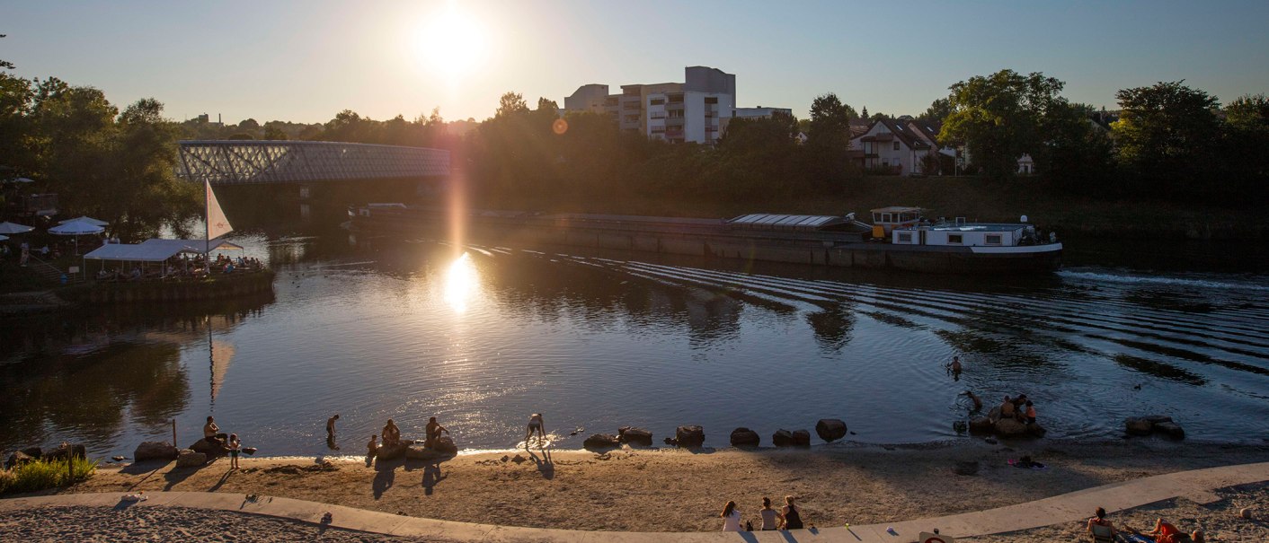 Sonnenuntergang am Neckarstrand in Remseck. Menschen entspannen am sandigen Ufer, während ein Schiff den Fluss passiert. Die Sonne spiegelt sich im Wasser., © Stuttgart-Marketing GmbH, Achim Mende Sonnenuntergang am Neckarstrand in Remseck. Menschen entspannen am sandigen Ufer, während ein Schiff den Fluss passiert. Die Sonne spiegelt sich im Wasser., © Stuttgart-Marketing GmbH, Achim Mende