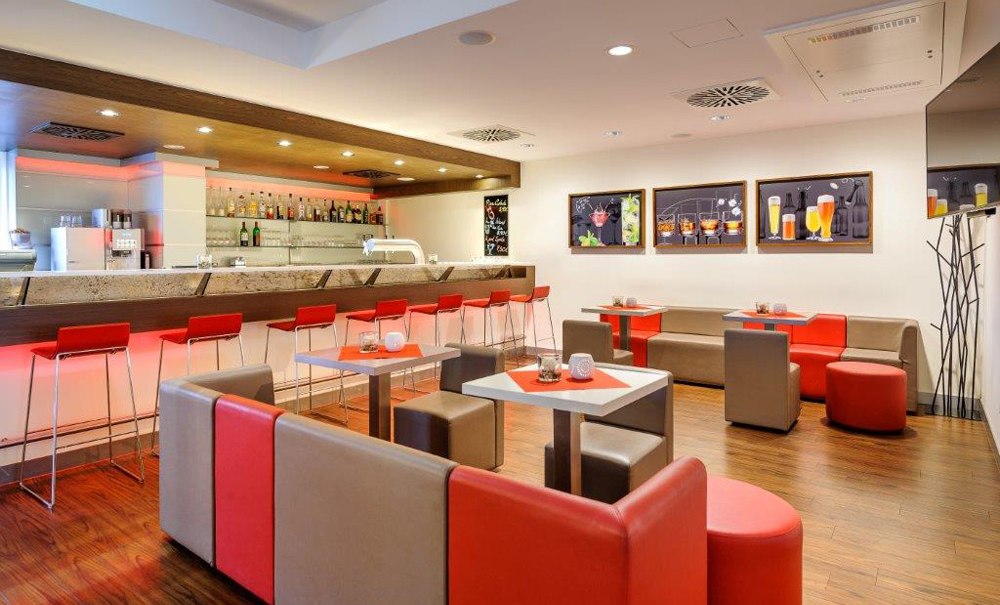 Moderne Bar mit roten und grauen Möbeln, beleuchtetem Tresen und Wandbildern von Getränken. Elegantes Ambiente mit Holzboden., © Ibis Stuttgart Airport Messe Moderne Bar mit roten und grauen Möbeln, beleuchtetem Tresen und Wandbildern von Getränken. Elegantes Ambiente mit Holzboden., © Ibis Stuttgart Airport Messe