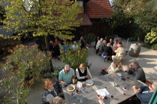 Gemütlicher Außenbereich mit mehreren Tischen, an denen Menschen essen und trinken. Umgeben von Pflanzen und Bäumen, entspannte Atmosphäre., © Elsbeth Koch