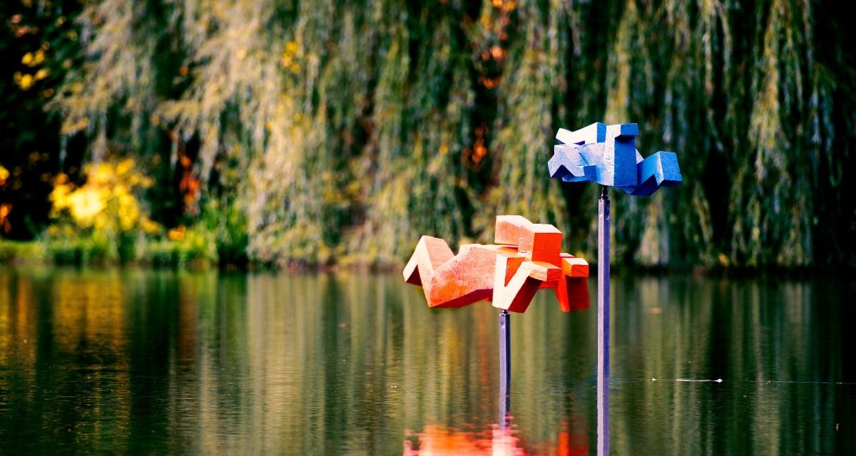 Zwei geometrische Skulpturen in Blau und Rot, die Schwimmer darstellen, stehen auf Stangen im Wasser eines Sees, umgeben von üppiger Vegetation., © Natur.Nah. Schönbuch & Heckengäu Zwei geometrische Skulpturen in Blau und Rot, die Schwimmer darstellen, stehen auf Stangen im Wasser eines Sees, umgeben von üppiger Vegetation., © Natur.Nah. Schönbuch & Heckengäu