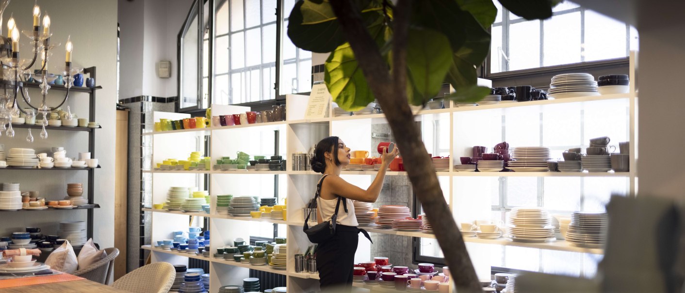 Frau in einem Geschäft betrachtet bunte Geschirrteile in Regalen. Der Raum ist hell erleuchtet, mit großen Fenstern und einem Kronleuchter., © SMG, Ingolf Pompe