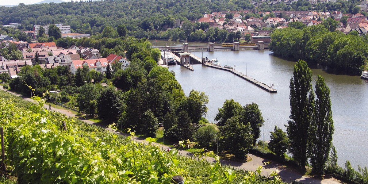 Blick auf den Neckar mit Schleuse, umgeben von Häusern, Bäumen und Weinbergen. Im Hintergrund eine bewaldete Hügellandschaft., © Stuttgart-Marketing GmbH Blick auf den Neckar mit Schleuse, umgeben von Häusern, Bäumen und Weinbergen. Im Hintergrund eine bewaldete Hügellandschaft., © Stuttgart-Marketing GmbH