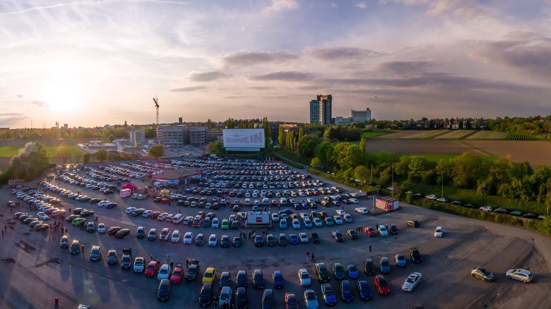 Luftaufnahme des Autokinos Kornwestheim bei Sonnenuntergang. Viele Autos sind geparkt, eine große Leinwand ist sichtbar. Im Hintergrund Felder und Gebäude., © DriveIn Autokinos DWJ GmbH
