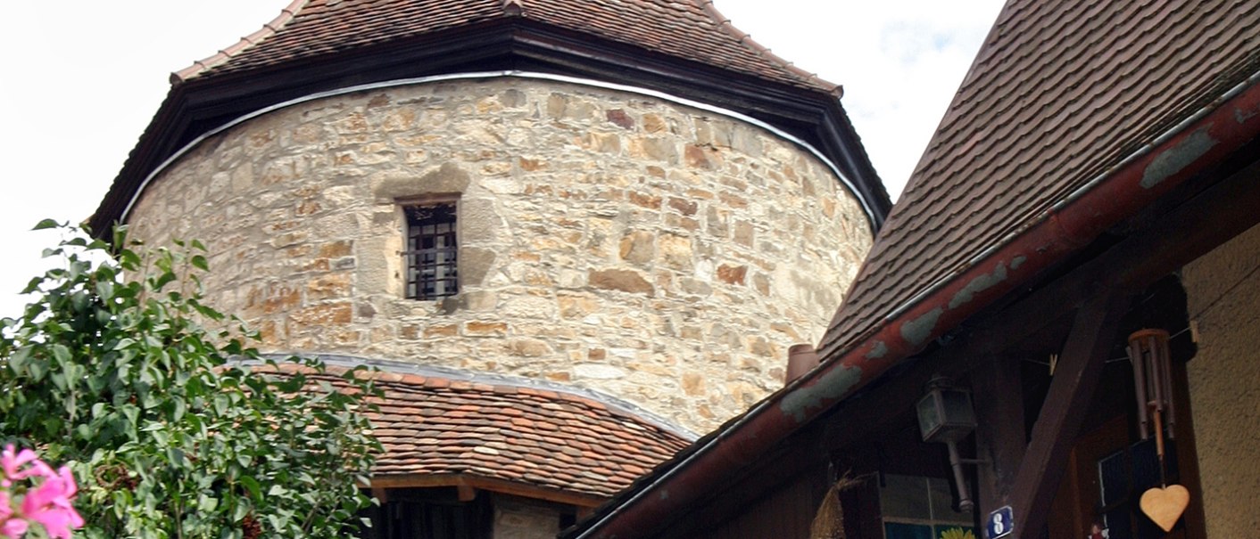 Ein mittelalterlicher Steinturm mit spitzem Dach erhebt sich über Fachwerkhäusern, umgeben von blühenden Blumen und blauem Himmel., © Mende Ein mittelalterlicher Steinturm mit spitzem Dach erhebt sich über Fachwerkhäusern, umgeben von blühenden Blumen und blauem Himmel., © Mende