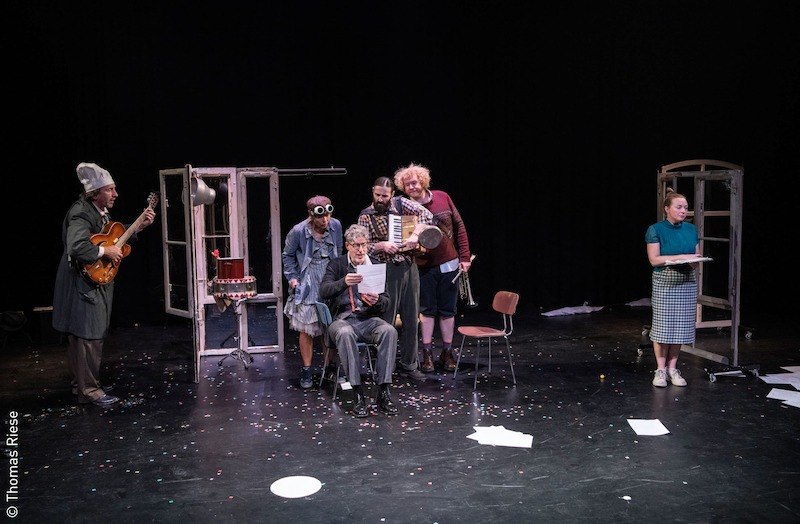 Theaterszene mit sechs Darstellern. Ein älterer Mann sitzt und liest, umgeben von Musikern und Schauspielern. Eine Frau steht rechts mit einem Tablett., © Stadt Kornwestheim Kulturmanagement