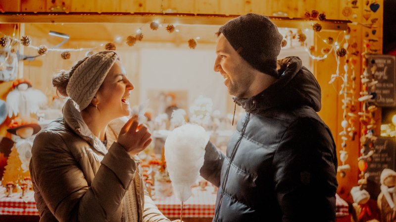 Ein Paar lacht und genießt Zuckerwatte vor einem festlich dekorierten Stand auf einem Weihnachtsmarkt., © Nik Ebert