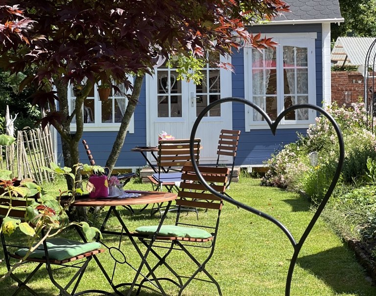 Garten mit Holztischen und -stühlen, umgeben von Pflanzen. Im Hintergrund ein blaues Häuschen und ein Baum mit roten Blättern., © Café Plätschwiesle