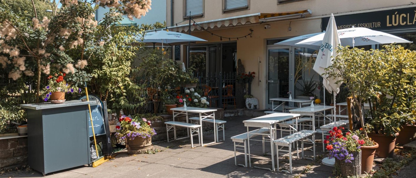 Gemütlicher Außenbereich eines Cafés mit weißen Tischen und Stühlen, umgeben von üppigen Pflanzen und bunten Blumen., © Stuttgart Marketing GmbH, Sarah Schmid Gemütlicher Außenbereich eines Cafés mit weißen Tischen und Stühlen, umgeben von üppigen Pflanzen und bunten Blumen., © Stuttgart Marketing GmbH, Sarah Schmid