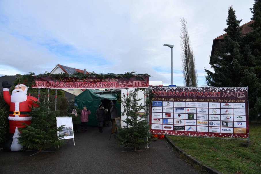 Eingang zum Weihnachtsmarkt Maitis mit einem großen Weihnachtsmann, Tannenbäumen und einem Banner mit Unterstützerlogos.