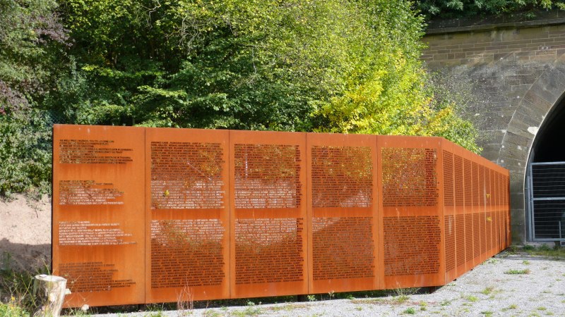 Die Namenswand an der KZ-Gedenkst&auml;tte Leonberg zeigt eingravierte Namen auf rostfarbenem Metall. Sie steht vor einem Tunnel, umgeben von gr&uuml;ner Vegetation., &copy; Stadt Leonberg