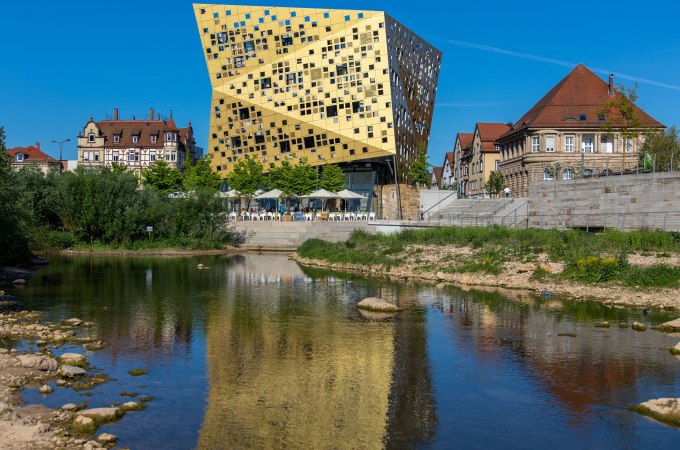 Ein modernes, goldenes Gebäude spiegelt sich in einem Fluss. Historische Gebäude und grüne Uferpflanzen umgeben die Szene unter klarem, blauem Himmel., © Touristik und Marketing GmbH