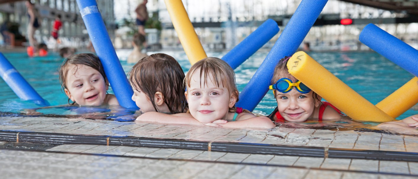 Vier Kinder im Schwimmbad lehnen am Beckenrand und halten bunte Schwimmnudeln. Sie lächeln und tragen Badeanzüge., © Badezentrum Sindelfingen