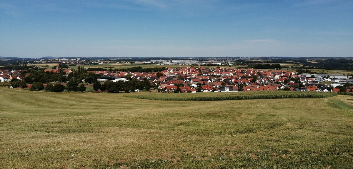Grüne Wiesen führen zu einer Siedlung mit roten Dächern, umgeben von Feldern und Wäldern unter blauem Himmel., © Foto: Cornelia Steinbach Grüne Wiesen führen zu einer Siedlung mit roten Dächern, umgeben von Feldern und Wäldern unter blauem Himmel., © Foto: Cornelia Steinbach