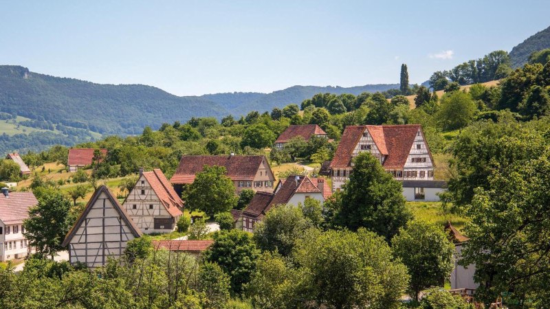 Ein malerisches Dorf mit traditionellen Fachwerkhäusern, umgeben von grünen Hügeln und Bäumen, unter einem klaren blauen Himmel., © SMG, Achim Mende Ein malerisches Dorf mit traditionellen Fachwerkhäusern, umgeben von grünen Hügeln und Bäumen, unter einem klaren blauen Himmel., © SMG, Achim Mende