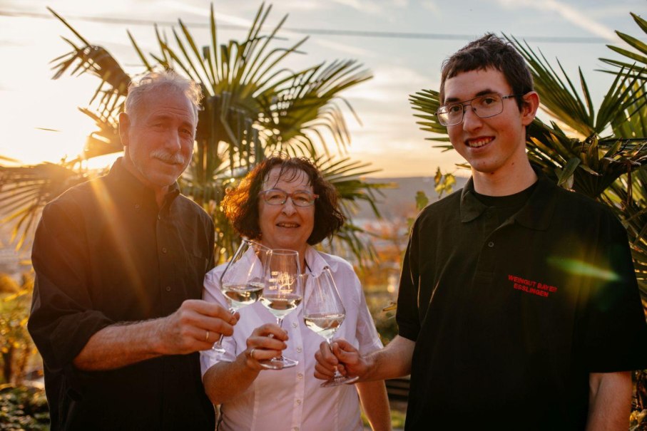 Drei Personen sto&szlig;en mit Weingl&auml;sern im Sonnenuntergang an. Sie stehen vor Palmen und wirken fr&ouml;hlich und entspannt., &copy; Weingut Bayer