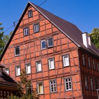 Ein gro&szlig;es Fachwerkhaus mit rotem Ziegelmauerwerk und dunklem Dach steht vor einem klaren blauen Himmel. B&auml;ume umgeben das Geb&auml;ude., &copy; Agentur Arcos
