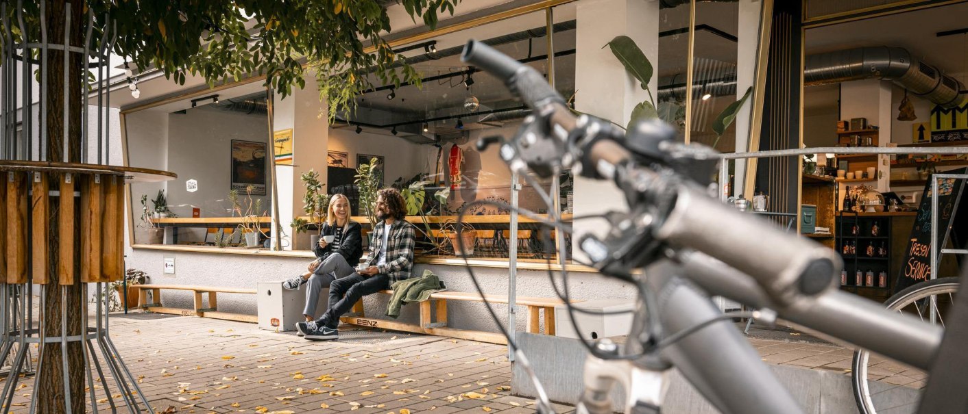 Zwei Personen sitzen lachend vor einem Caf&eacute; auf einer Bank. Im Vordergrund ist ein Fahrradlenker zu sehen. Herbstliche Bl&auml;tter liegen auf dem Boden., &copy; SMG Stuttgart Marketing GmbH - Sarah Schmid
