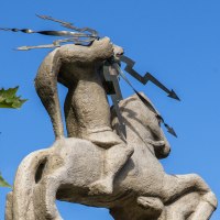 Steinfigur eines Reiters mit Blitzsymbolen, umgeben von grünen Blättern, vor klarem, blauem Himmel., © SMG, Achim Mende