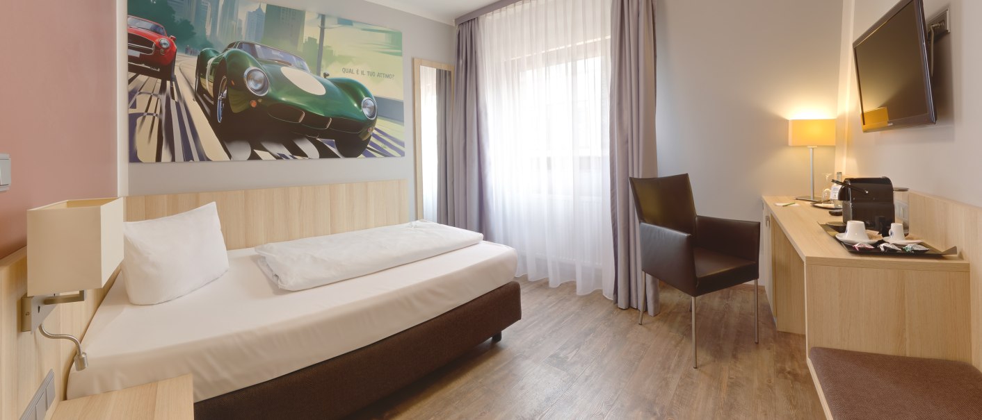 Modernes Einzelzimmer mit Bett, Schreibtisch, Stuhl und großem Wandbild von Autos. Helle Vorhänge und Holzfußboden schaffen eine einladende Atmosphäre., © attimo Hotel Stuttgart