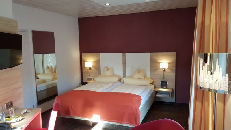 Modernes Doppelzimmer mit zwei Betten, roter Wand, Fernseher und stilvoller Beleuchtung. Ein Tisch mit Wasserflasche und Gläsern steht bereit., © Hotel Koch