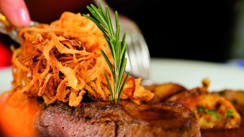 Zwiebelrostbraten mit knusprigen Zwiebeln und Rosmarin garniert. Im Hintergrund sind Beilagen zu sehen., © Kzenon-Fotolia