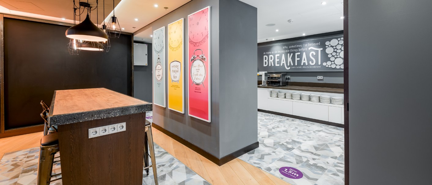 Moderner Frühstücksbereich mit Holztisch, Hockern und dekorativen Postern. Im Hintergrund ein Buffetbereich mit Geschirr und Frühstücksangebot., © Premier Inn