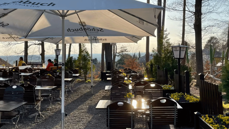 Ein ruhiger Biergarten mit Holztischen und St&uuml;hlen unter Sonnenschirmen, umgeben von B&auml;umen. Einige G&auml;ste sitzen und genie&szlig;en die Aussicht., &copy; Sch&ouml;nbuch Waldbiergarten am Wasserturm