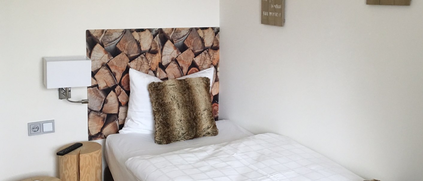 Ein gemütliches Einzelbett mit Holzoptik-Kopfteil, Fellkissen und Decke. An der Wand hängen motivierende Sprüche., © TOMAS Ein gemütliches Einzelbett mit Holzoptik-Kopfteil, Fellkissen und Decke. An der Wand hängen motivierende Sprüche., © TOMAS
