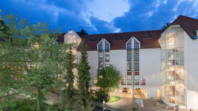 Das Best Western Plus Hotel am Schlossberg bei D&auml;mmerung, mit beleuchteten Fenstern und einem gl&auml;sernen Treppenhaus, umgeben von B&auml;umen., &copy; Best Western Plus Hotel am Schlossberg