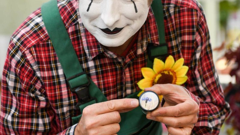 Ein Pantomime mit weißem Gesicht und Strohhut zeigt auf eine Lupe mit Blume. Er trägt ein kariertes Hemd und grüne Hosenträger.