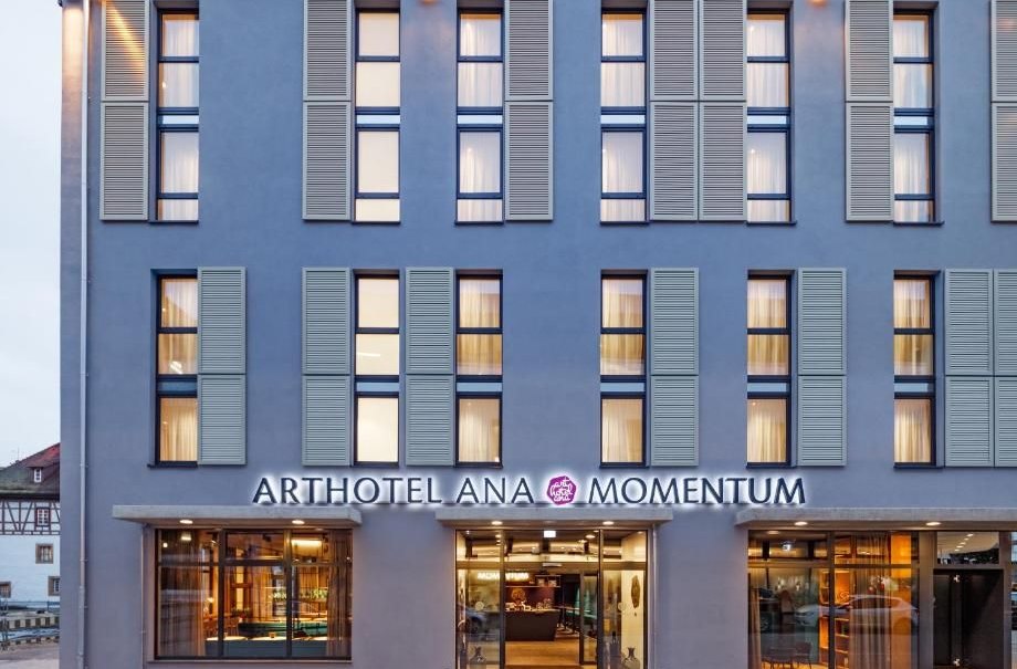 Fassade des Arthotel Ana Momentum mit großen Fenstern und beleuchtetem Eingang. Moderne Architektur mit blauen Fensterläden., © Arthotel ANA Momentum