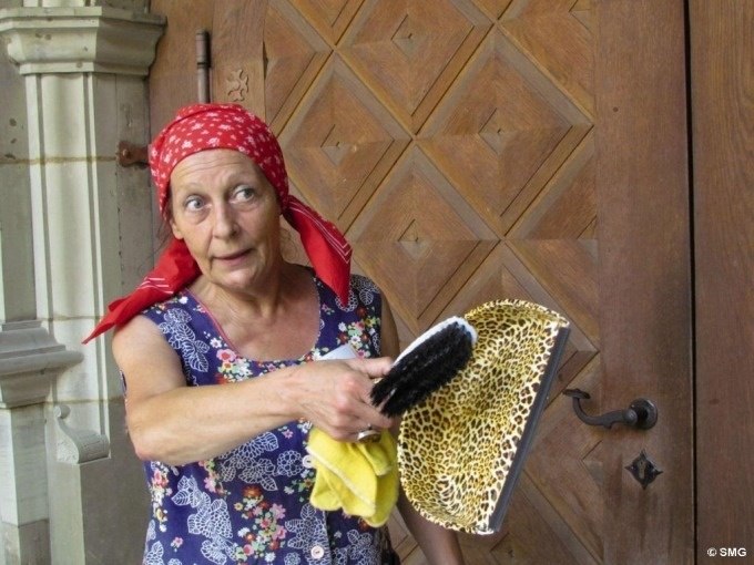 Eine Frau mit rotem Kopftuch hält Putzutensilien vor einer verzierten Holztür. Sie trägt ein buntes Kleid und schaut aufmerksam., © Stuttgart Marketing GmbH Eine Frau mit rotem Kopftuch hält Putzutensilien vor einer verzierten Holztür. Sie trägt ein buntes Kleid und schaut aufmerksam., © Stuttgart Marketing GmbH