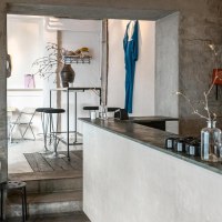 Modernes Café mit minimalistischer Einrichtung, weißen Wänden und Holzmöbeln. Ein blaues Kleidungsstück hängt an der Wand, Pflanzen auf Tischen., © SMG, Sarah Schmid