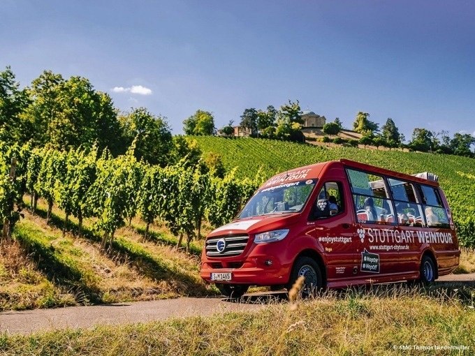 Ein roter Tourbus mit der Aufschrift 'Stuttgart Weintour' fährt durch grüne Weinberge unter blauem Himmel., © SMG, Thomas Niedermüller Ein roter Tourbus mit der Aufschrift 'Stuttgart Weintour' fährt durch grüne Weinberge unter blauem Himmel., © SMG, Thomas Niedermüller