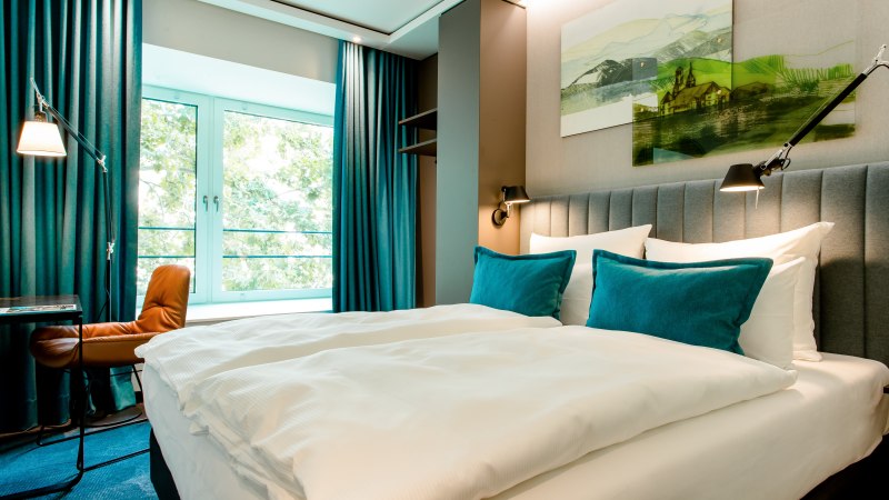 Modernes Hotelzimmer mit großem Bett, blauen Kissen und Vorhängen. Ein Fenster bietet Blick ins Grüne. Ein Schreibtisch mit Stuhl steht daneben., © Motel One - Stuttgart Hauptbahnhof