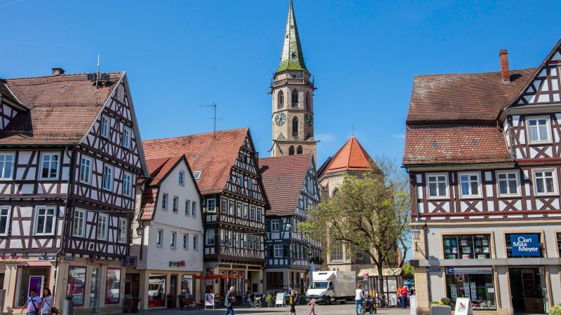 Fachwerkhäuser am Marktplatz in Schorndorf, mit einem Kirchturm im Hintergrund. Menschen spazieren über den Platz bei sonnigem Wetter., © Stuttgart-Marketing GmbH