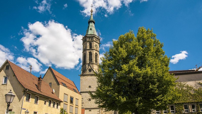 Die Amanduskirche in Bad Urach erhebt sich majestätisch neben Wohnhäusern unter einem klaren blauen Himmel mit weißen Wolken., © Stuttgart-Marketing GmbH, Sarah Schmid