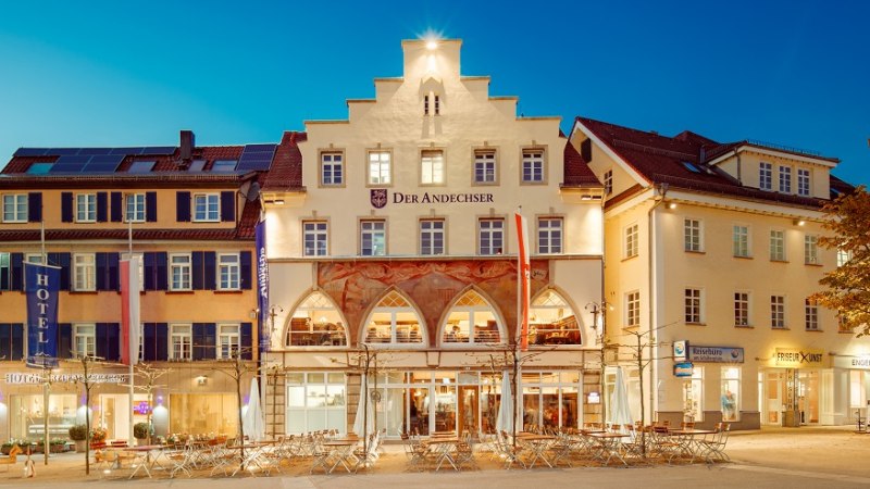 Abendansicht von „Der Andechser“ mit Hotel, beleuchteten Fenstern und Außengastronomie an einem Platz., © Tobias Fröhner Abendansicht von „Der Andechser“ mit Hotel, beleuchteten Fenstern und Außengastronomie an einem Platz., © Tobias Fröhner