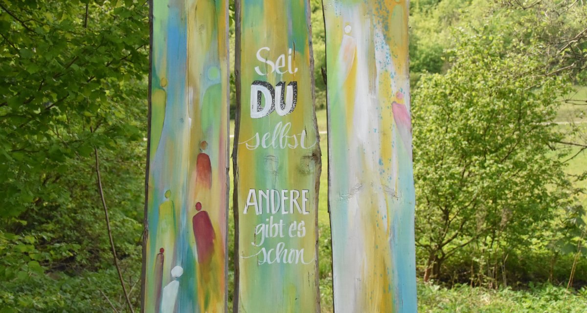 Bunte Holzstele im Wald mit dem Spruch 'Sei du selbst, andere gibt es schon'. Umgeben von grüner Natur., © Ev. Kirchenbezirk Schorndorf Bunte Holzstele im Wald mit dem Spruch 'Sei du selbst, andere gibt es schon'. Umgeben von grüner Natur., © Ev. Kirchenbezirk Schorndorf