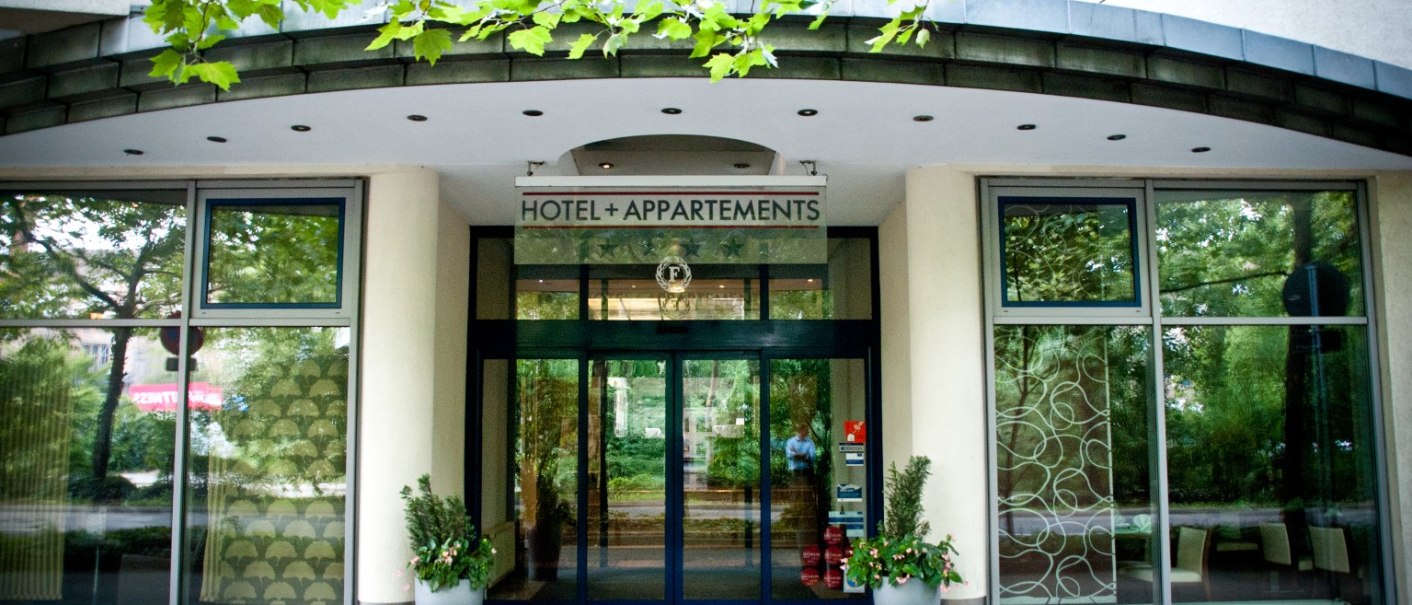 Hoteleingang mit Glasfront und der Aufschrift 'Hotel + Appartements'. Zwei große Pflanzkübel flankieren den Eingang., © City Hotel Reutlingen