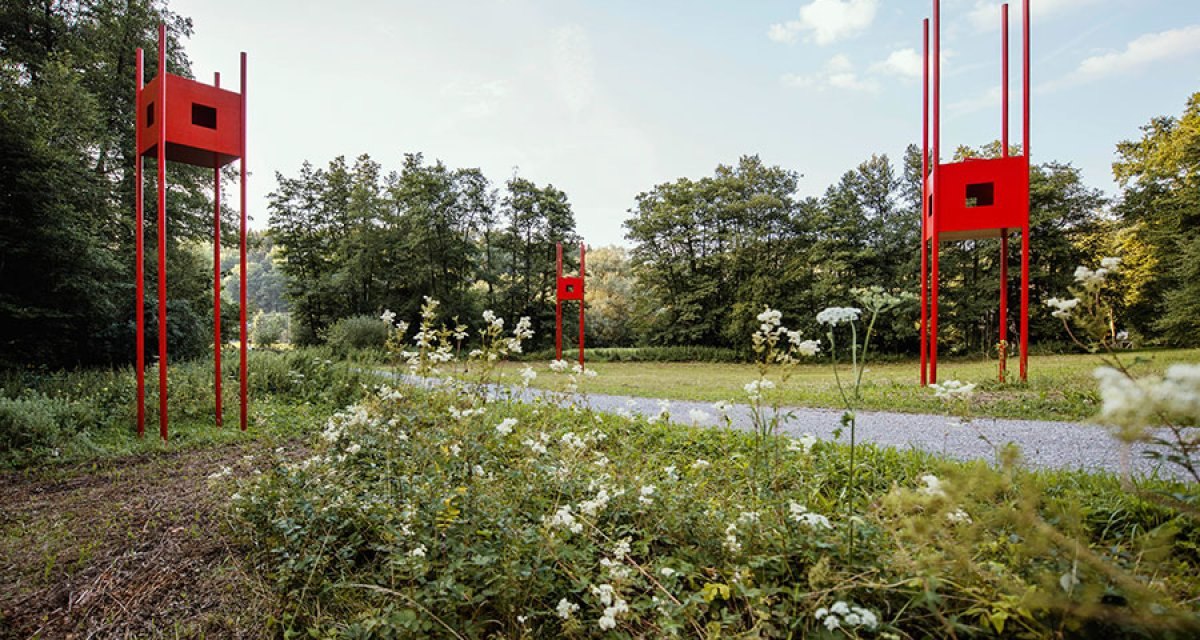 Rote Skulpturen mit quadratischen Elementen stehen auf hohen Stangen in einer Waldlandschaft. Weiße Blumen blühen im Vordergrund., © Natur.Nah. Schönbuch & Heckengäu Rote Skulpturen mit quadratischen Elementen stehen auf hohen Stangen in einer Waldlandschaft. Weiße Blumen blühen im Vordergrund., © Natur.Nah. Schönbuch & Heckengäu