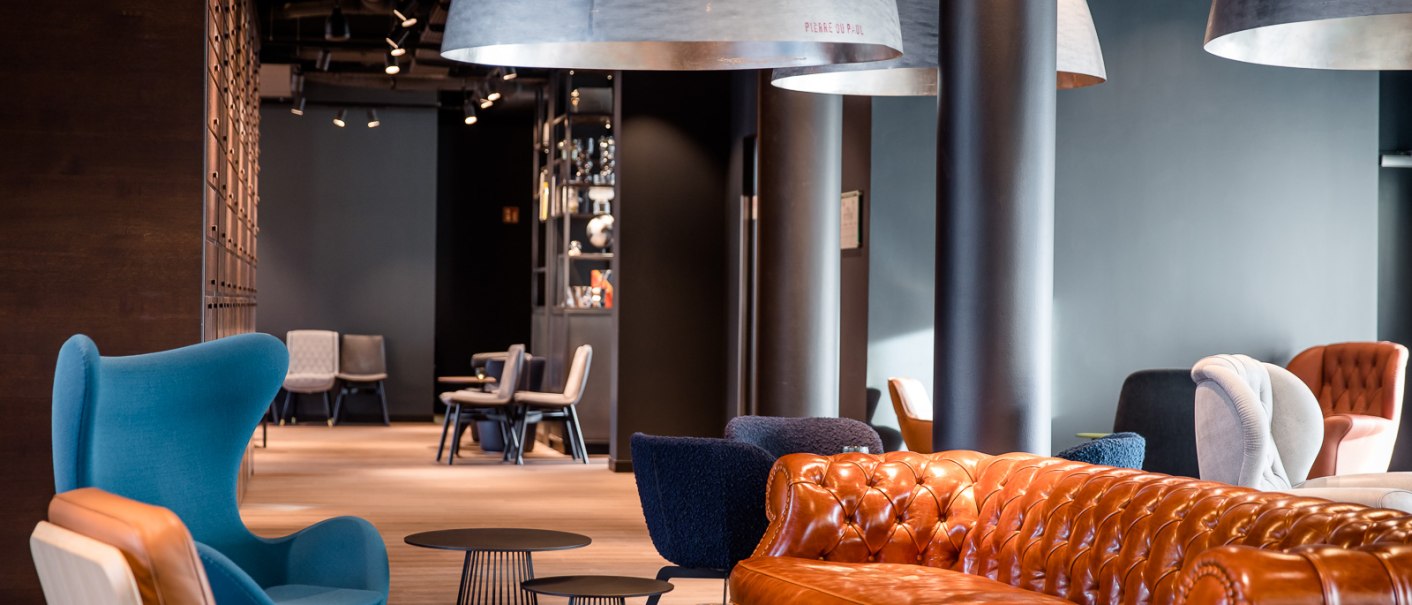 Moderne Lounge mit braunem Ledersofa, blauen und grauen Sesseln, großen Hängelampen und Holzboden. Im Hintergrund sind weitere Sitzgelegenheiten sichtbar., © TOMAS Moderne Lounge mit braunem Ledersofa, blauen und grauen Sesseln, großen Hängelampen und Holzboden. Im Hintergrund sind weitere Sitzgelegenheiten sichtbar., © TOMAS